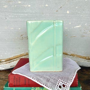 May include: A rectangular, light green ceramic vase with a glossy finish. The vase has a geometric design with diagonal lines and is displayed on top of a stack of books and a white lace doily.