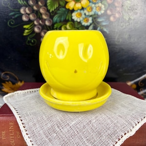 May include: A bright yellow ceramic planter with a smiling face design. The planter sits on a matching saucer. The cheerful design adds a playful touch to any space. The planter is likely for small plants or succulents.