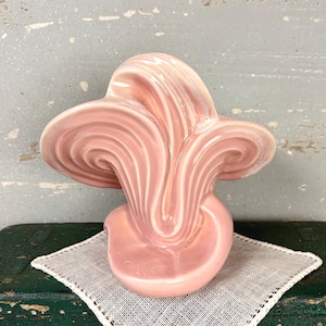 May include: A glossy, light pink ceramic sculpture featuring a series of fluid, wave-like ridges that fan outward from a central point. The object has a smooth, reflective finish and a curved, organic shape that creates a sense of movement.