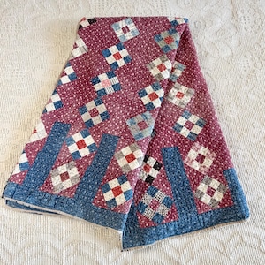 May include: A folded quilt featuring a deep maroon background with a small white floral pattern. The quilt is decorated with a repeating grid of square blocks in shades of blue, white, and red, along with solid blue rectangular border accents.