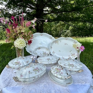 May include: A set of vintage white porcelain serving dishes with a delicate floral pattern in shades of pink and green. The dishes are arranged on a white tablecloth with lace trim, and are surrounded by a bouquet of pink and white flowers.