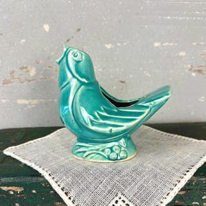 May include: A glossy teal ceramic bird figurine with a hollow center, featuring molded wing details and a base decorated with small circular berry-like shapes. The bird is depicted with its head tilted upward and its beak slightly open.