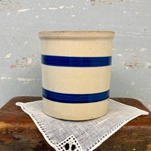 May include: A cylindrical ceramic crock featuring a light beige, speckled finish with two prominent, horizontal dark blue stripes encircling the body. The vessel has a flat rim and a sturdy, straight-walled design with a smooth, matte texture.