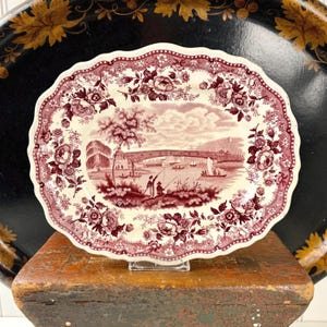 May include: An oval ceramic platter with a scalloped edge featuring a maroon transferware pattern. The center depicts a landscape with a bridge, boats, and figures fishing, while the border is decorated with detailed floral motifs.