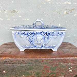 May include: A blue and white ceramic trinket box with a lid. The box features a raised relief design with a cameo portrait and floral motifs. The box has a handle on the lid and sits on small feet.