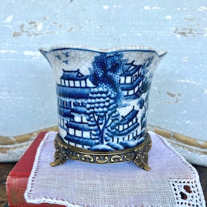 May include: A blue and white ceramic planter with a detailed Asian-inspired landscape design. The planter sits on a decorative brass stand. The rim of the planter has a scalloped edge.
