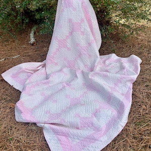May include: A pink and white quilt with a geometric pattern. The quilt is draped over a surface, showcasing its design and texture. The quilt's edges are bordered with a solid pink fabric.