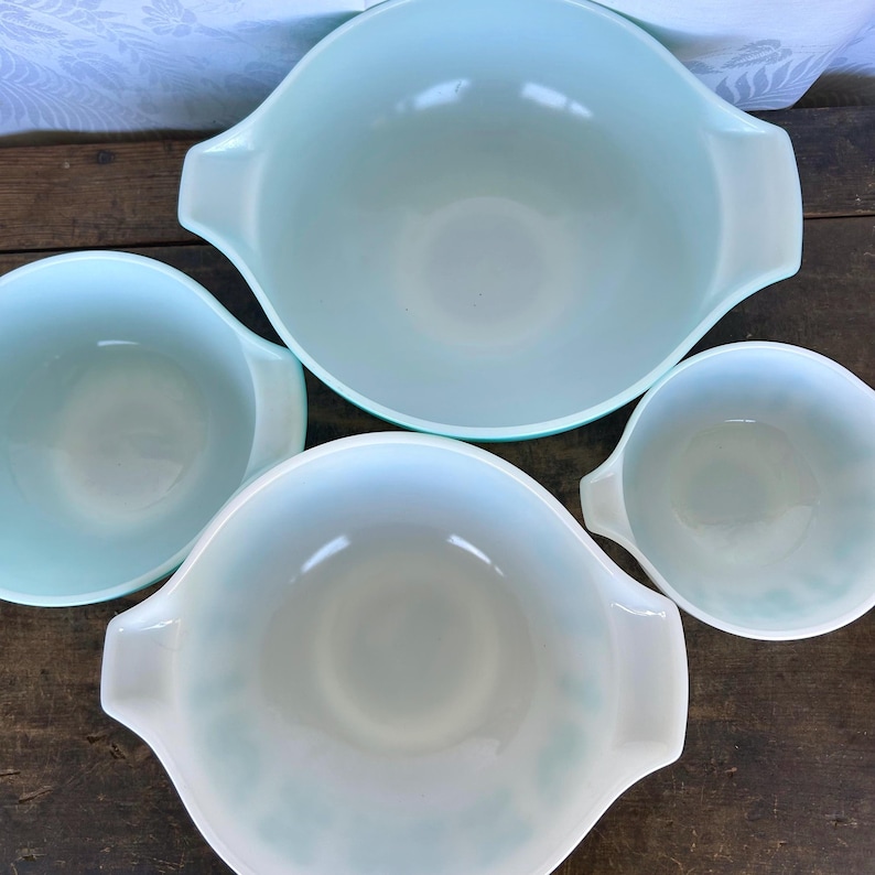 May include: Set of four vintage blue mixing bowls with a white interior. The bowls are nested together, with the largest bowl on top and the smallest bowl on the bottom.