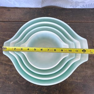 May include: Set of four nesting mixing bowls in a light blue color. The bowls are made of glass and have a smooth, rounded shape. A measuring tape is placed across the bowls, showing the largest bowl is 12 inches in diameter.