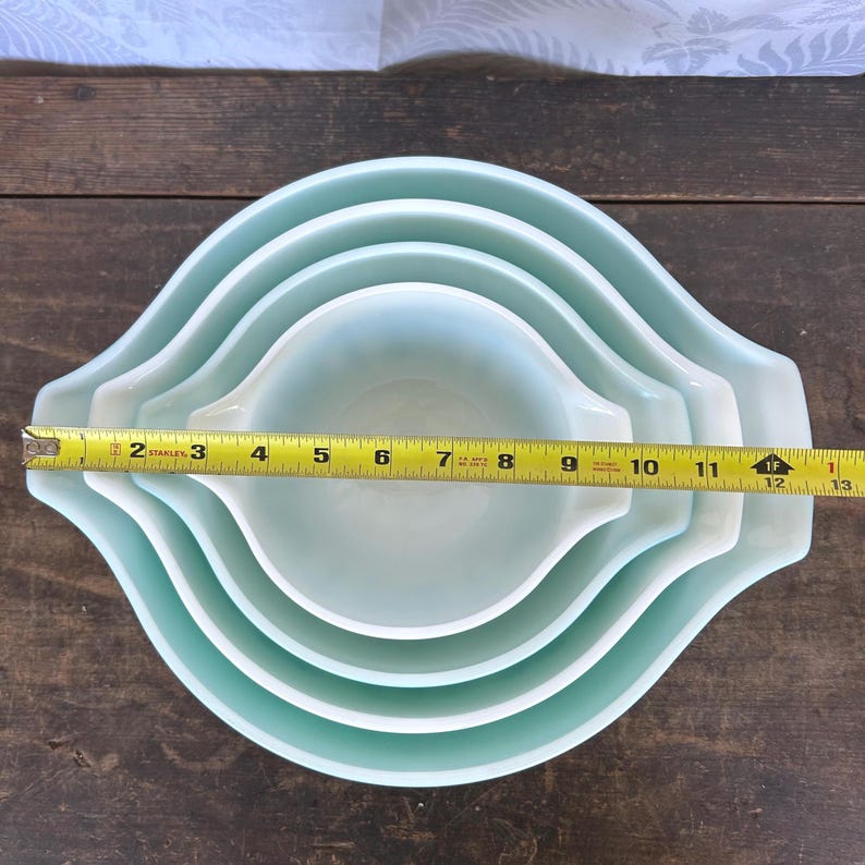 May include: Set of four nesting mixing bowls in a light blue color. The bowls are made of glass and have a smooth, rounded shape. A measuring tape is placed across the bowls, showing the largest bowl is 12 inches in diameter.