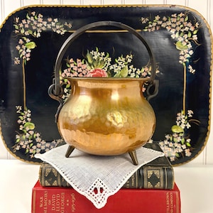 May include: A hammered copper pot with a dark metal swing handle and three small feet. The pot sits on a white lace doily atop two stacked books, one red and one black, in front of a large, dark blue decorative tray with floral patterns.