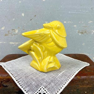 May include: A bright yellow ceramic figurine of a bird perched on a stylized base. The bird has a smooth, glossy finish with molded wing and feather details. The sculpture is depicted in a side profile view, showcasing its beak and rounded head.