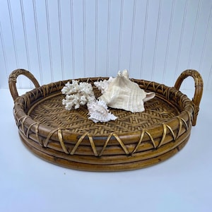 May include: A round, woven bamboo tray with two curved handles. The tray is filled with a white coral and two seashells. The tray has a natural brown color with gold accents and is suitable for serving or display.