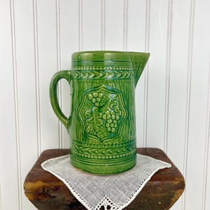 May include: A green ceramic pitcher with a handle and spout. The pitcher features a raised grape design in the center and decorative patterns around the top and bottom. It sits on a small wooden surface with a white cloth.