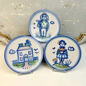 May include: Three ceramic plates with hand-painted designs. Each plate features a white background with a blue border. The designs include a person with a rake, a house, and a person in a dress. The plates are displayed on clear stands.