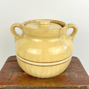 May include: A vintage, yellow-glazed ceramic pot with a lid. The pot has two handles and a decorative band around the middle. The lid has a knob and a ridged design. The pot is likely used for cooking or storage.