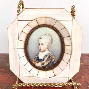 May include: A miniature portrait of a young person in a white cap and blue and gold dress, framed in a white and gold octagonal frame with a gold stand.