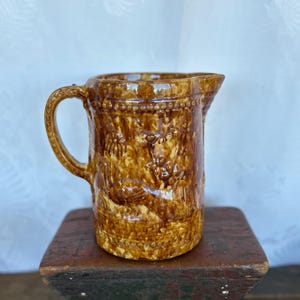 May include: A brown and yellow ceramic pitcher with a floral design. The pitcher has a handle and a rounded base.