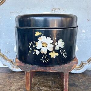 May include: A black metal container with a lid, featuring a floral design in white and yellow. The container has white handles and sits on a small wooden stool. The floral pattern is on the front of the container.
