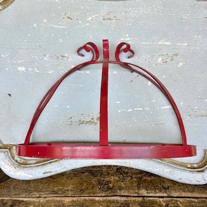 May include: Red metal wall-mounted shelf with a curved top and a flat base. The shelf has a decorative design at the top and is likely used for displaying items. The shelf is painted a bright red color.
