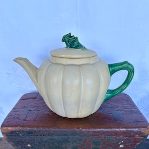May include: A cream-colored ceramic teapot with a green pumpkin-shaped lid and handle. The teapot has a ribbed design and is sitting on a wooden surface.