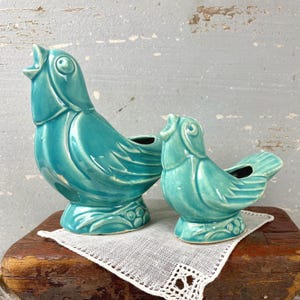 May include: Two turquoise ceramic bird-shaped planters with glossy finishes. Each bird is depicted with an open beak, textured wing details, and a base featuring molded berry or leaf patterns. The larger bird stands to the left of the smaller bird.