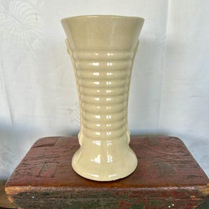 May include: A cream-colored ceramic vase with a ribbed design and a flared opening. The vase has a smooth, glossy finish.