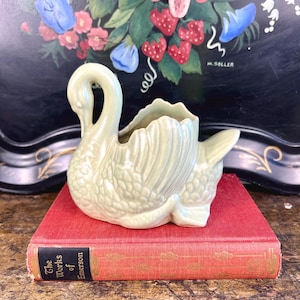 May include: A pale green ceramic swan planter with a textured surface. The swan's head is turned to the side and its wings are spread out to form a planter. The planter is sitting on a red book with the title "The Works of Emerson".