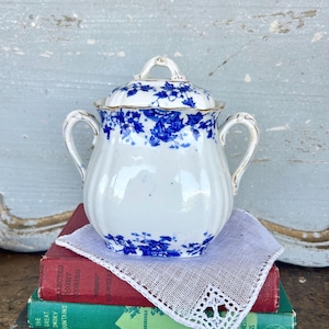 May include: White porcelain sugar bowl with a lid, adorned with blue floral patterns. The bowl has two handles and a gold trim. It sits on a white lace doily, resting on top of a stack of books.