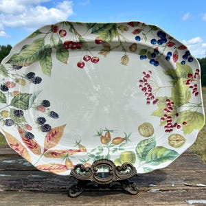 May include: A white oval serving platter with a decorative border of green leaves, red berries, and blackberries. The platter is on a wooden stand.