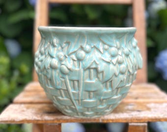 Pode incluir: Um vaso de cerâmica turquesa com um design de tecido de cesta e detalhes florais e de bagas em relevo. O vaso redondo está sobre uma cadeira de madeira, com um fundo desfocado de flores azuis e vegetação.