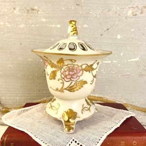 May include: A white porcelain decorative container with a lid, featuring a pink floral design and gold accents. The container has a pedestal base with three gold feet and a gold handle on the lid. The lid has several small holes.