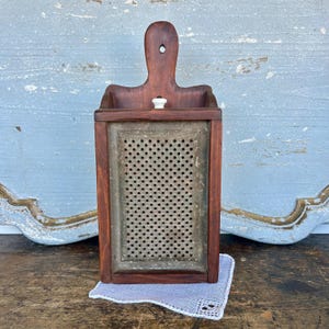 May include: A vintage wooden grater with a dark brown frame and handle. The grater has a metal grating surface and a small white knob. It sits on a white lace doily, with a light blue background.