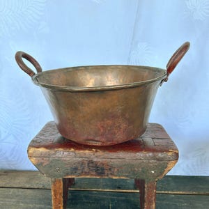 May include: A vintage copper bowl with two wooden handles. The bowl is sitting on a wooden stool.