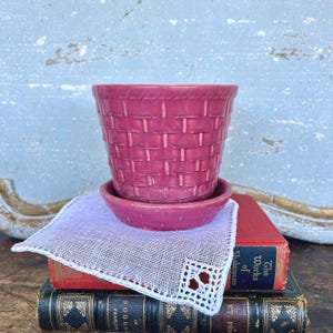 May include: A magenta ceramic planter with a basket weave design, sitting on a matching saucer. The planter and saucer are placed on a stack of vintage books and a white lace-edged handkerchief.