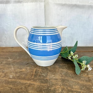 May include: A blue and white striped ceramic pitcher with a curved handle and spout. The pitcher has a light blue body with horizontal white stripes. The handle and spout are off-white. A sprig of greenery with small white flowers is next to the pitcher.