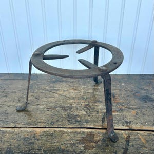 May include: A rustic, three-legged metal trivet with a circular top. The trivet is made of dark, aged metal with visible wear and tear, suggesting it is an antique. The legs are slender and angled, providing stability. The design is simple and functional.