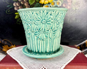 May include: A turquoise ceramic plant pot with a matching saucer. The pot has a repeating daisy pattern and vertical ribbed detailing. The pot is on a white lace doily and a red book.
