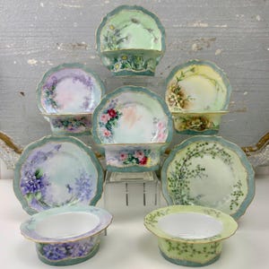 May include: A collection of antique porcelain dishes with scalloped edges and floral designs. Each dish features a unique hand-painted floral arrangement in pastel colors, including daisies, roses, and violets, with a teal border and gold trim.