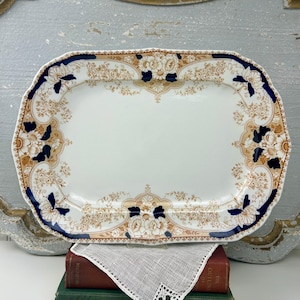 May include: Rectangular antique serving platter with a white center and a decorative border. The border features floral patterns in shades of brown, tan, and navy blue. The platter has a scalloped edge and is likely made of ceramic.