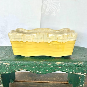 May include: A yellow and white ceramic planter with a wavy rim and a glazed finish. The planter is sitting on a green wooden stool.