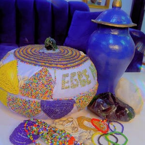 Puede incluir: Un recipiente blanco redondeado decorado con cuentas de colores, conchas de cauri y el texto "EGBE" en cuentas doradas. Junto a él hay una jarra azul con tapa, una piedra oscura translúcida, una piedra blanca y collares de cuentas.
