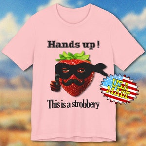 May include: Pink t-shirt with a humorous graphic of a strawberry wearing a black mask and mustache, holding a gun. The text "Hands up!" and "This is a strobbery" are printed above and below the strawberry. A "USA MADE" sticker is on the right.