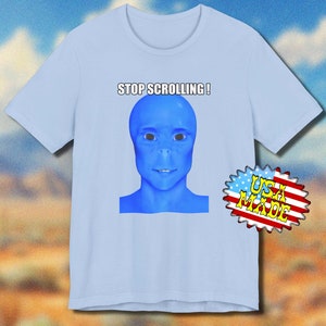 Può includere: T-shirt azzurra con un volto alieno blu e la scritta "STOP SCROLLING!". La maglietta ha maniche corte e girocollo. Un adesivo "USA MADE" è visibile sul lato destro.