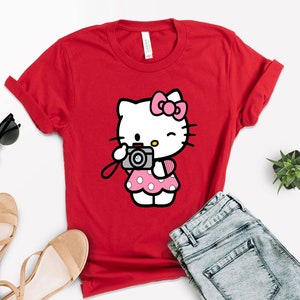 Pode incluir: Camiseta vermelha com um gr&aacute;fico branco de Hello Kitty. Hello Kitty est&aacute; segurando uma c&acirc;mera rosa e preta e vestindo um vestido rosa com bolinhas brancas.