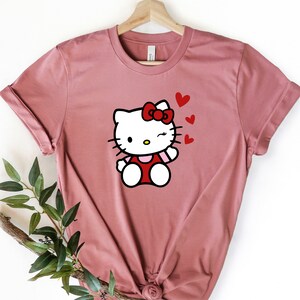 Op de afbeelding: Een roze T-shirt met een witte Hello Kitty-afbeelding die een rode jurk en een rode strik draagt. Hello Kitty knippert met haar oog en heeft rode hartjes om haar heen.