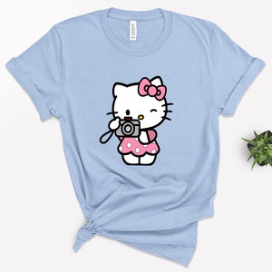 Pode incluir: Uma camiseta azul claro com uma imagem de desenho animado do Hello Kitty segurando uma c&acirc;mera. Hello Kitty est&aacute; vestindo um vestido rosa com bolinhas brancas e um la&ccedil;o rosa.