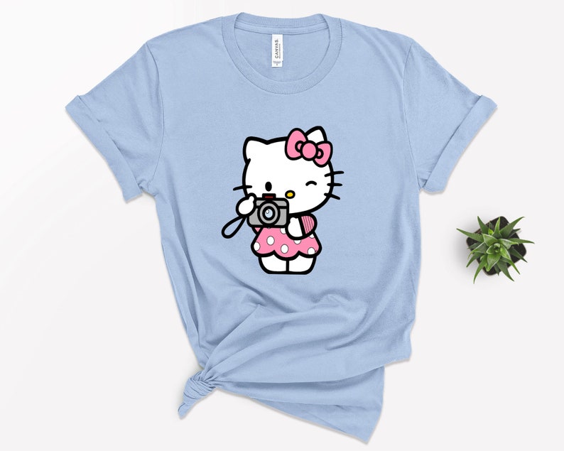 Pode incluir: Uma camiseta azul claro com uma imagem de desenho animado do Hello Kitty segurando uma c&acirc;mera. Hello Kitty est&aacute; vestindo um vestido rosa com bolinhas brancas e um la&ccedil;o rosa.