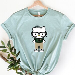 Puede incluir: Una camiseta azul claro con una imagen de dibujos animados de Hello Kitty con gafas y una camisa a cuadros.