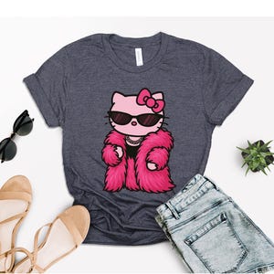 Puede incluir: Camiseta gris oscuro con un gato de dibujos animados que lleva gafas de sol y un abrigo de piel rosa. El gato tiene un lazo rosa y un collar. La camiseta se combina con pantalones cortos de mezclilla, gafas de sol y sandalias.